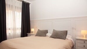 A neatly made bed with a beige blanket, two bedside tables with lamps, and a window with sheer curtains. - Casa Centro Torrevieja San Policarpo (Torrevieja)