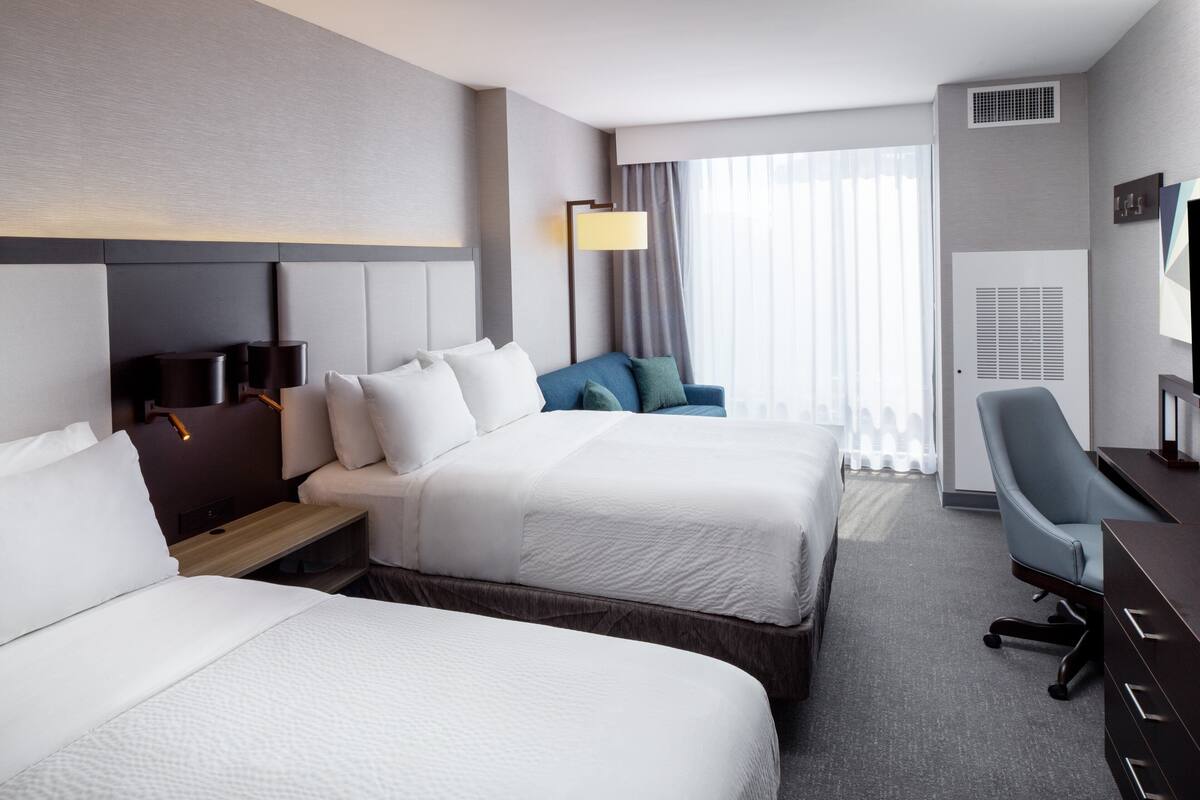 A hotel room with two beds, a desk, a chair, a lamp, and a large window with curtains.