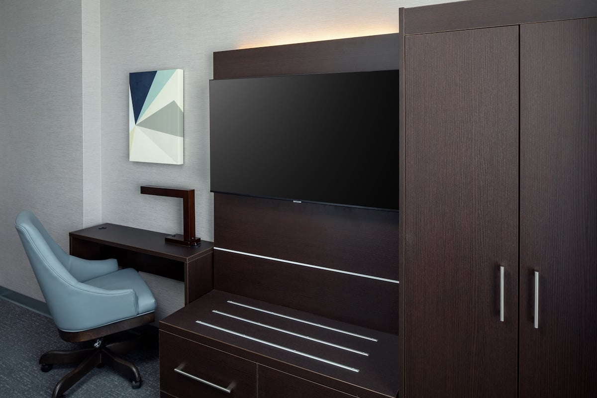 A modern hotel room with a dark wood desk, a light blue office chair, a flat-screen TV, and a cabinet with drawers.