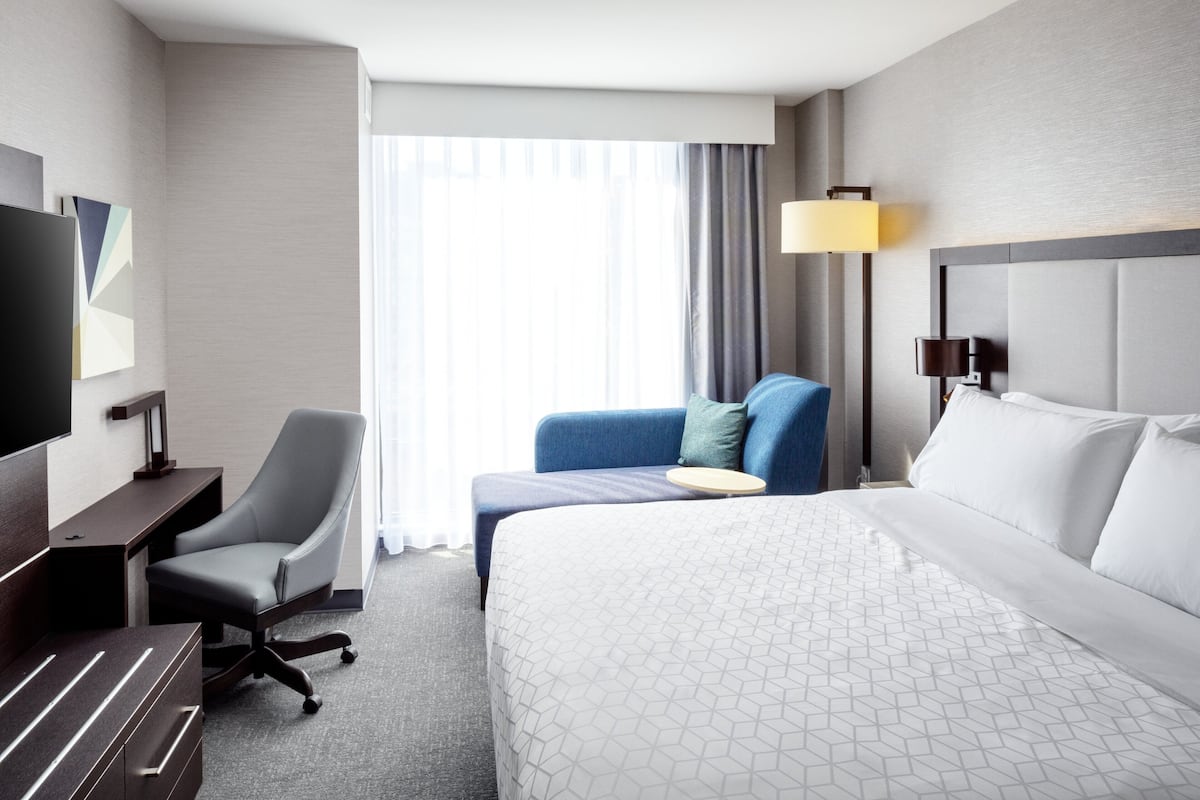 A hotel room with a large bed, a desk, a chair, a TV, and a window with curtains.
