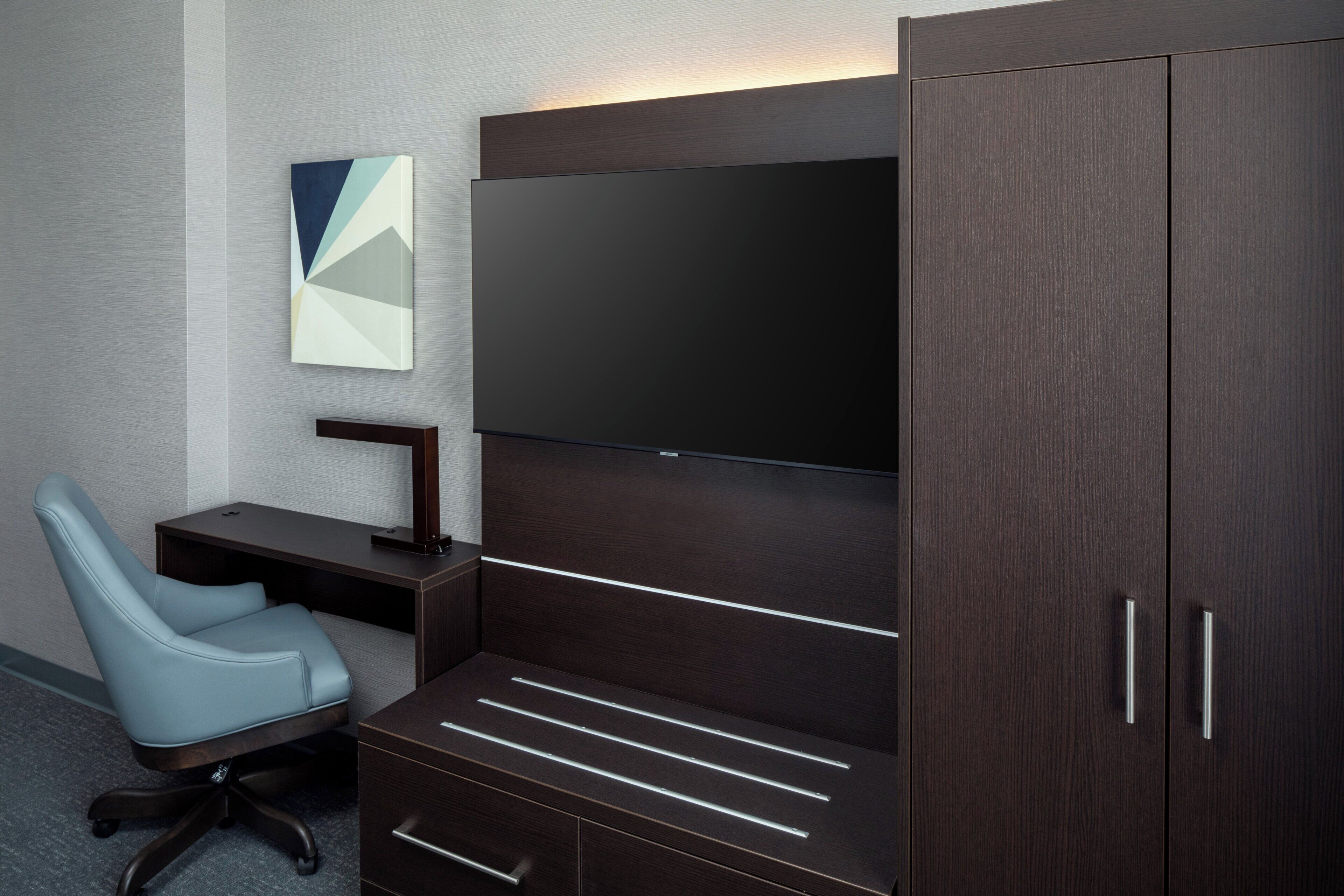 A modern office space with a desk, chair, and a large flat-screen TV mounted on a dark wood cabinet.