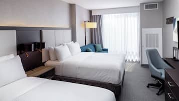 Habitación de hotel con dos camas, un escritorio, una silla, una lámpara y un ventanal grande con cortinas.
