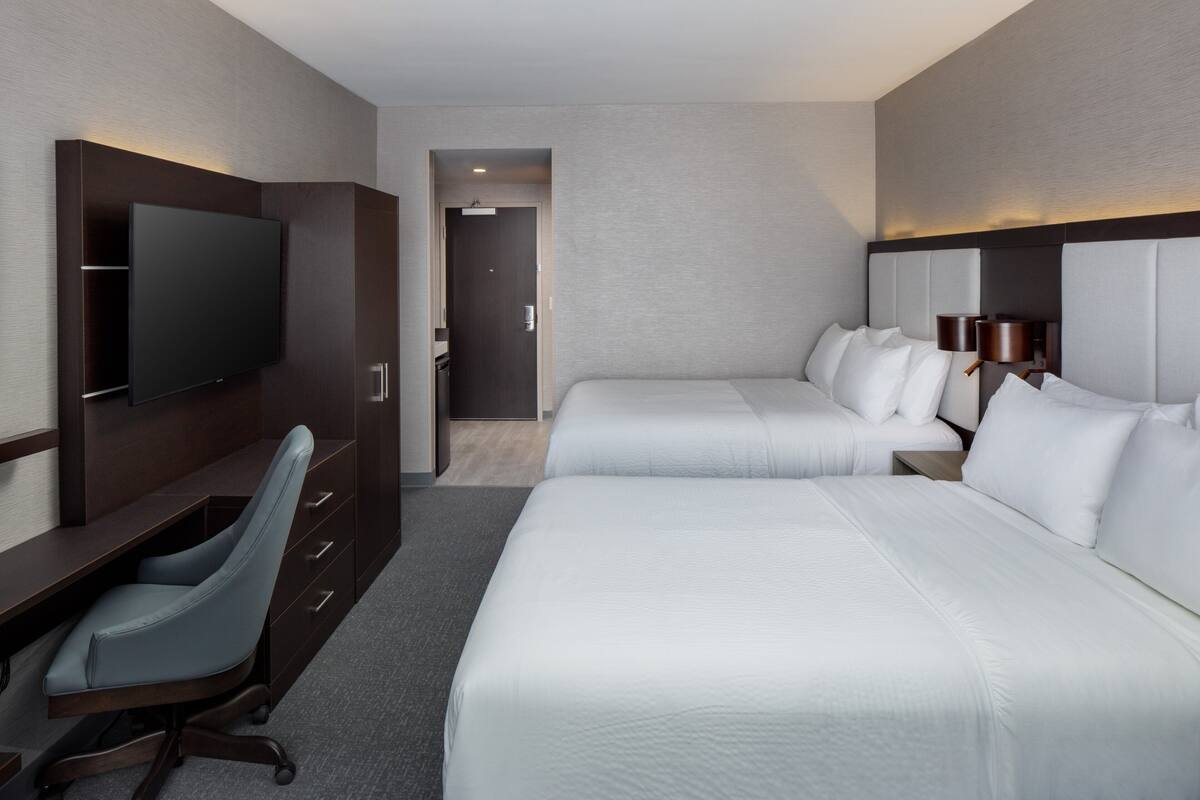 A hotel room with a flat-screen TV, a desk with a chair, two beds with headboards, and a lamp.
