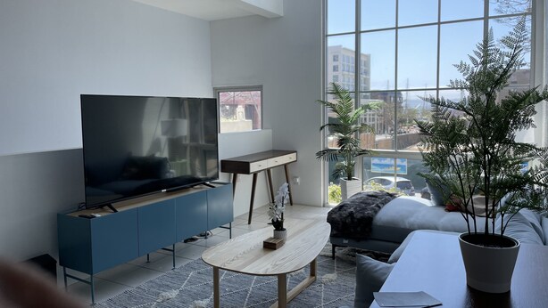 Uma sala de estar moderna com uma televisão grande, um armário azul, uma mesa de centro redonda, um sofá cinza e uma janela ampla com vista para a cidade.