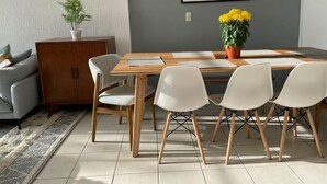 Uma sala de jantar com mesa de madeira e quatro cadeiras brancas, um sofá cinzento, um armário e um vaso com flores amarelas.
