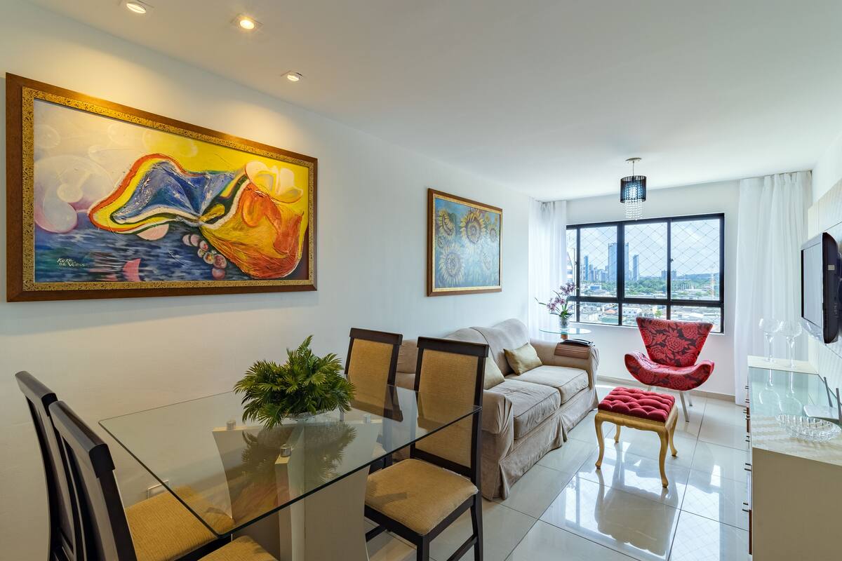 A modern living room with a glass dining table, beige sofa, red armchair, and large paintings on the walls.