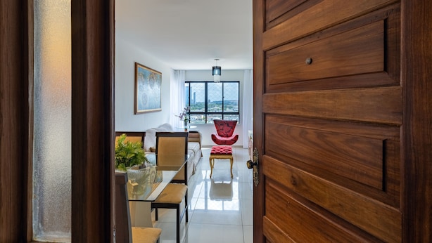 A modern interior with a wooden door marked "903", a dining area with a glass table, chairs, and a red chair, and a view through a large window. - Excelente comodidade em Recife (Recife)