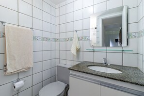 Un baño con paredes de azulejos blancos, un inodoro, un lavabo con encimera de granito y un espejo con repisa de vidrio.