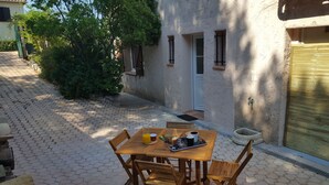 Ein Holztisch mit Stühlen und einem Tablett mit Frühstücksangeboten, auf einer gepflasterten Terrasse mit einem Haus im Hintergrund.
