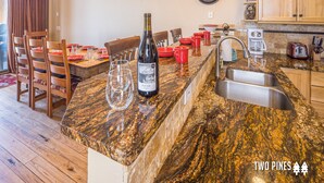 Une cuisine dotée de plans de travail en granit, d’une bouteille de vin et d’un coin repas avec des chaises en bois et de la vaisselle rouge.