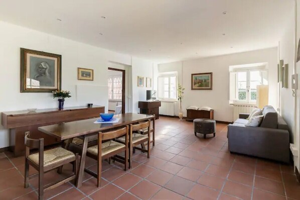 A spacious living area with a wooden dining table, a sofa, and a TV. There are framed pictures on the walls and a window providing natural light. - Renovated Grange in a Renaissance Villa Park (Capannori)