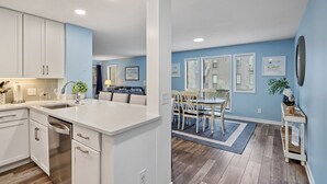 Une cuisine moderne avec des meubles blancs et un îlot central, un coin repas agrémenté d’un tapis bleu et de chaises en bois, et un salon avec un mur bleu et une grande fenêtre.