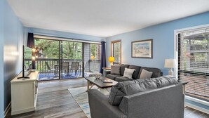 Un salon moderne avec un canapé gris, une table basse en bois et un téléviseur posé sur un meuble. Il y a un balcon avec vue sur les arbres et une rambarde.