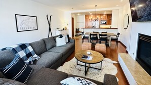 Een moderne woonkamer met een bank, salontafel en een open haard. Er is een eethoek met een tafel en stoelen. De keuken is op de achtergrond zichtbaar, met houten kasten en een kroonluchter.