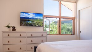 Een slaapkamer met een bed, een dressoir met lades, een aan de muur bevestigde televisie en een groot raam met uitzicht op een bos en bergen.