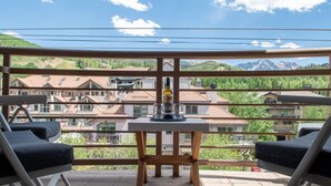 Een balkon met twee loungestoelen, een tafel met een fles en glazen, en uitzicht op huizen en bergen.