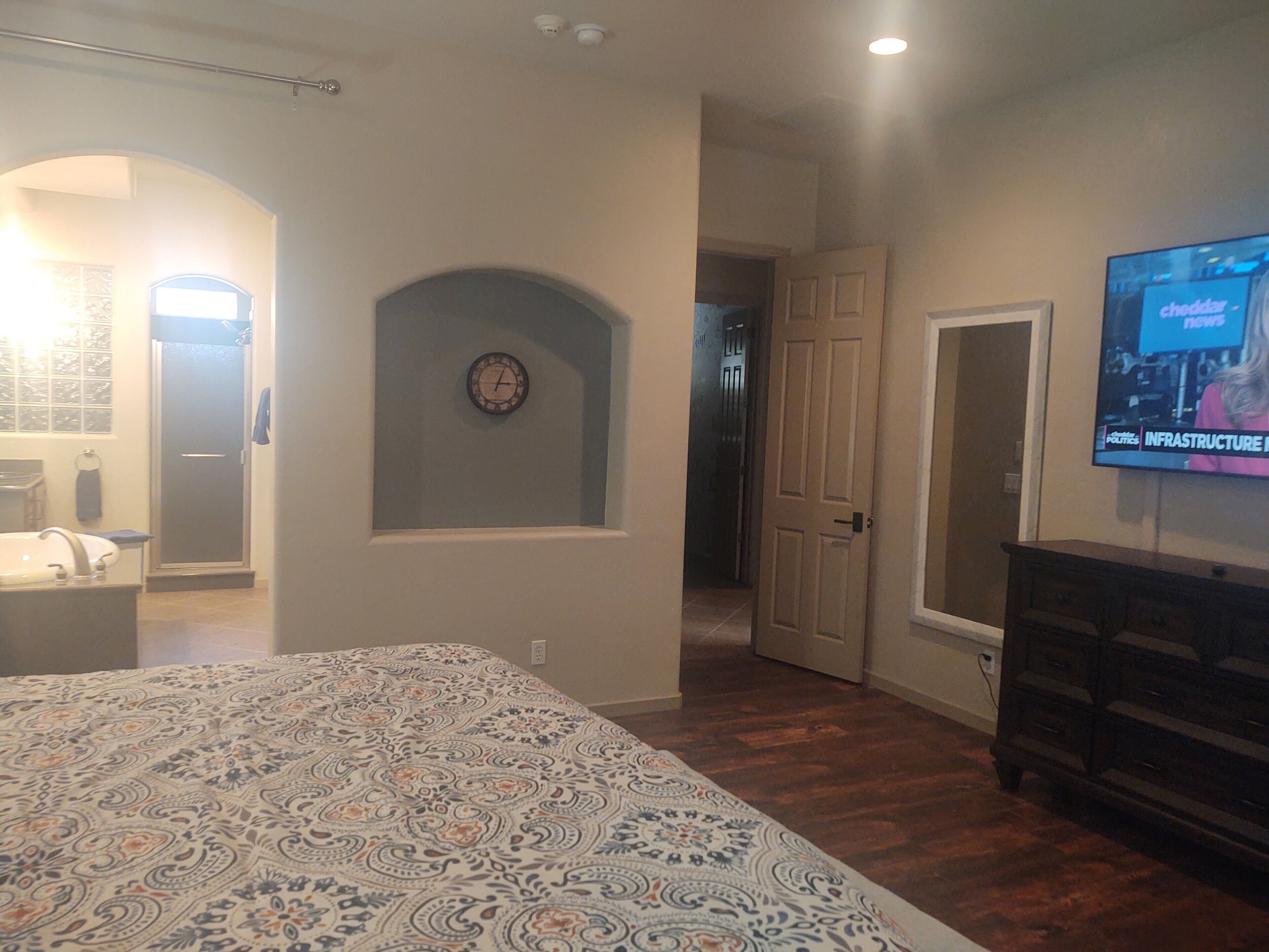 A bedroom with a bed, a TV, a clock, and a hallway leading to a bathroom.