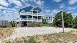 Ein zweistöckiges Strandhaus mit Balkon, vor dem sich ein Sandstrand und ein Volleyballnetz befinden.