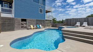 Ein Swimmingpool mit blau gefliestem Rand, umgeben von einer Betonplattform. Weiße und blaue Liegestühle und ein Holzzaun sind im Hintergrund zu sehen.
