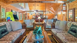 A cozy cabin interior with wooden walls and ceilings, two sofas with floral upholstery, a central coffee table with a decorative plant, and a fireplace with a stone surround. - Now and Forever (Sevierville)