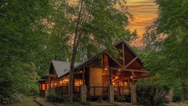 A log cabin with a stone foundation, wooden exterior, and a porch with railings. - Now and Forever (Sevierville)