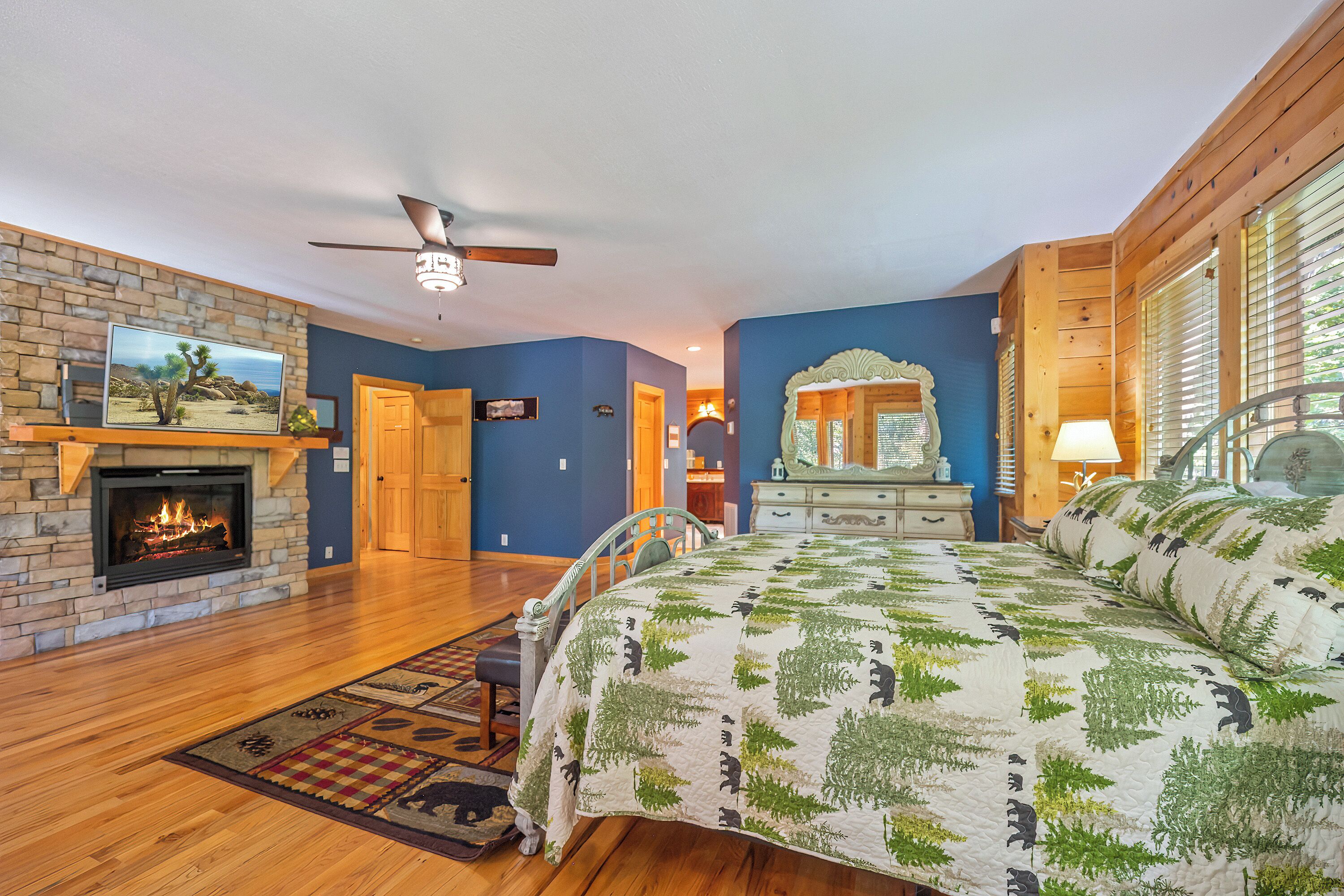 A bedroom with a stone fireplace, a bed with a forest-themed quilt, a ceiling fan, and a large mirror.