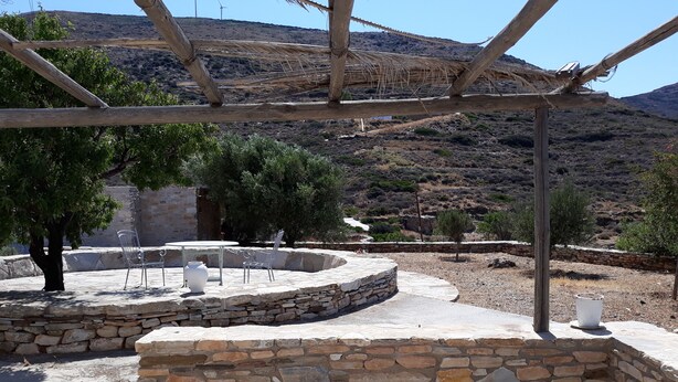 Zona salotto all'aperto con muri in pietra e sedie in metallo, circondata da alberi e un paesaggio montuoso.