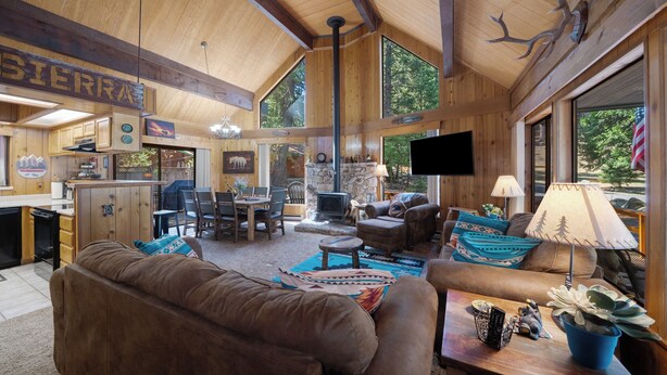 A spacious living area with a fireplace, wooden furniture, and large windows offering a view of the outdoors. - Woodsy Mountain Cabin - Best Location In Graeagle (Graeagle)