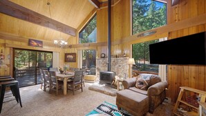 A spacious living room with a stone fireplace, wooden furniture, and large windows offering a view of the forest. - Woodsy Mountain Cabin - Best Location In Graeagle (Graeagle)