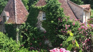 Une maison traditionnelle, dotée d’un toit à forte pente, entourée d’une végétation luxuriante et de fleurs en pleine floraison.