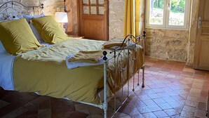 Une chambre à coucher rustique avec un mur en pierre, une porte en bois, une fenêtre ornée de rideaux jaunes et un cadre de lit en métal.