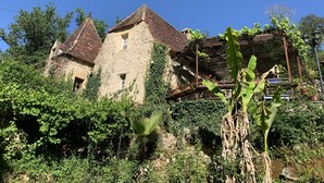 Un bâtiment rustique en pierre, doté d’un toit en tuiles, entouré d’une végétation luxuriante et traversé par un petit ruisseau.