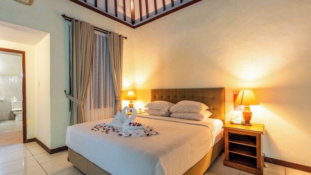 Ein Hotelzimmer mit einem großen Bett, zwei Nachttischlampen, einem Holznachttisch und Blick ins Badezimmer.