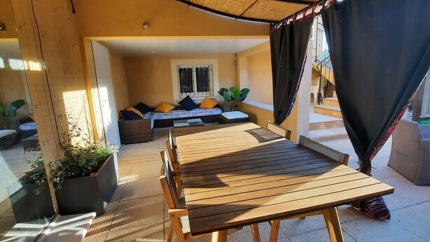 Un espace extérieur aménagé, abrité, avec une table et des chaises en bois, un canapé avec des coussins et des plantes en pot.