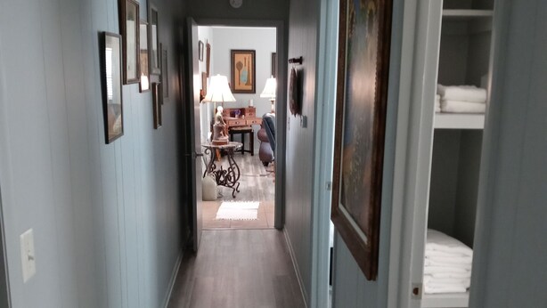 A hallway with light blue walls, wooden flooring, and framed pictures on the walls. There is a door on the right leading to a room with a bed. - Cozy Hideaway in Town (Springdale)