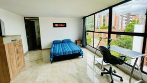 Une chambre à coucher avec un lit, un bureau et une vue sur les immeubles, grâce à de grandes fenêtres.