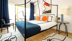 A four-poster bed with a blue and white bedspread, orange pillows, and a painting of a flamingo on the wall. - Bright apartment steps from Praia da Rocha, one of Portugal’s finest beaches. Access to a large pool, promenade, restaurants and cafés. Sleeps 5. Self check-in via lockbox. (Portimão)