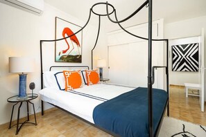 Ein Bett mit Baldachin, mit blauer Bettdecke und orangefarbenen Kissen, eine blaue Lampe auf einem Nachttisch und ein Flamingo-Gemälde an der Wand.