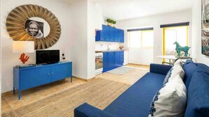 A compact living space with a blue sofa, a television on a blue cabinet, a kitchen area with blue cabinets, and a large mirror with a sunburst design. - Bright apartment steps from Praia da Rocha, one of Portugal’s finest beaches. Access to a large pool, promenade, restaurants and cafés. Sleeps 5. Self check-in via lockbox. (Portimão)