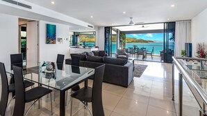 A modern living room with a glass dining table, dark sofas, and a view of the sea.