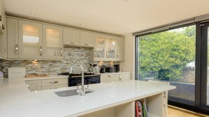 A modern kitchen with a large island, built-in appliances, and a view of greenery through a sliding glass door. - Fidra Cottage - Stunning Place to Stay! (North Berwick)