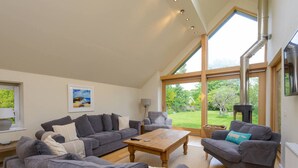 A spacious living room with grey sofas, a wooden coffee table, and large windows offering a view of a garden and a chimney. - Fidra Cottage - Stunning Place to Stay! (North Berwick)