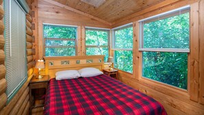 Un dormitorio tipo cabaña de madera con una cama, dos lámparas de noche y ventanas amplias que ofrecen vista a la vegetación.
