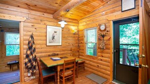 Interior acogedor de una cabaña con paredes de madera, un comedor con mesa y sillas, un cuadro en la pared, un reloj y una puerta con vistas a la vegetación exterior.