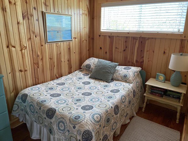 A bedroom with a wooden paneled wall, a bed with patterned bedding, a window with blinds, a bedside table with a lamp and books, and a small rug.