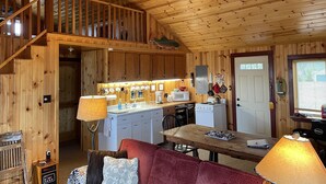 Interior de una cabaña de madera con cocina, comedor y una escalera que sube a un nivel superior.