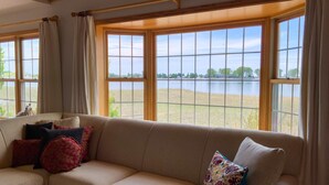 Una sala de estar con un sofá seccional color beige, varios cojines y ventanas amplias que ofrecen vistas a un lago y árboles.