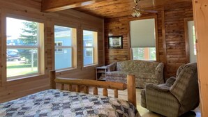 A log cabin bedroom with a bed, two chairs, a ceiling fan, and large windows. - Paul's Place - Two blocks from beaches in the heart of Grand Marais (Grand Marais)