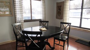 A dining area with a round table and four chairs, large windows with curtains, and a framed map on the wall. - Aspen House - Stunning 4 Bedroom A-Frame (Grand Marais)