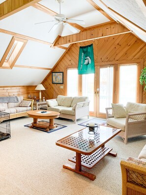 A cozy, well-lit room with wooden walls and a sloped ceiling, featuring a sofa set, a coffee table, and a hanging Spartan's Will flag.